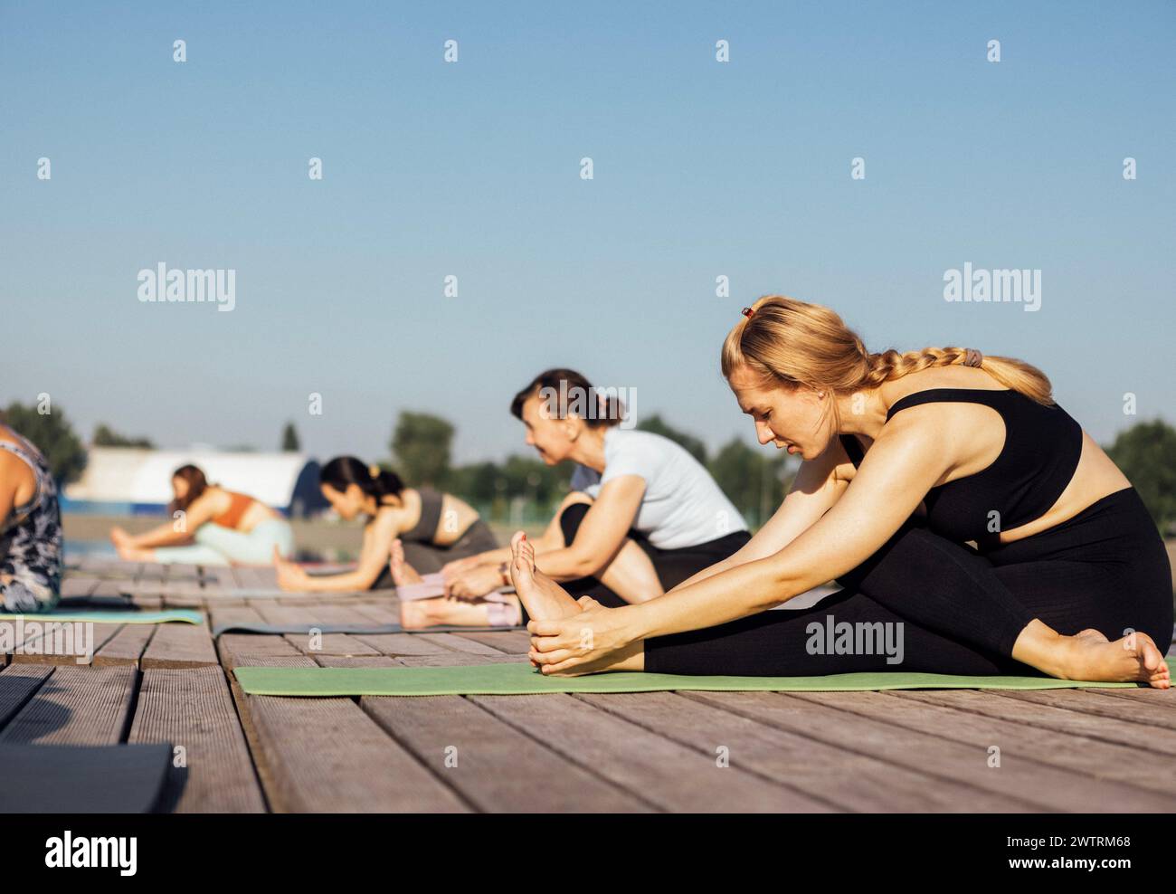 Lezione di yoga mattutina con istruttore vicino al fiume in città. Le giovani donne si piegano mentre si siedono sui tappetini in un molo di legno. Un gruppo di ragazze multietniche sono in agguato Foto Stock