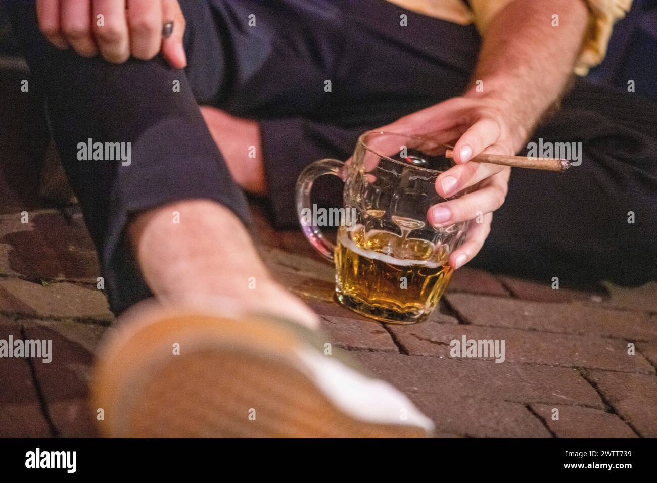 Una vista ravvicinata della mano di una persona che tiene un sigaro con una tazza di birra appoggiata su un pavimento in mattoni. Foto Stock