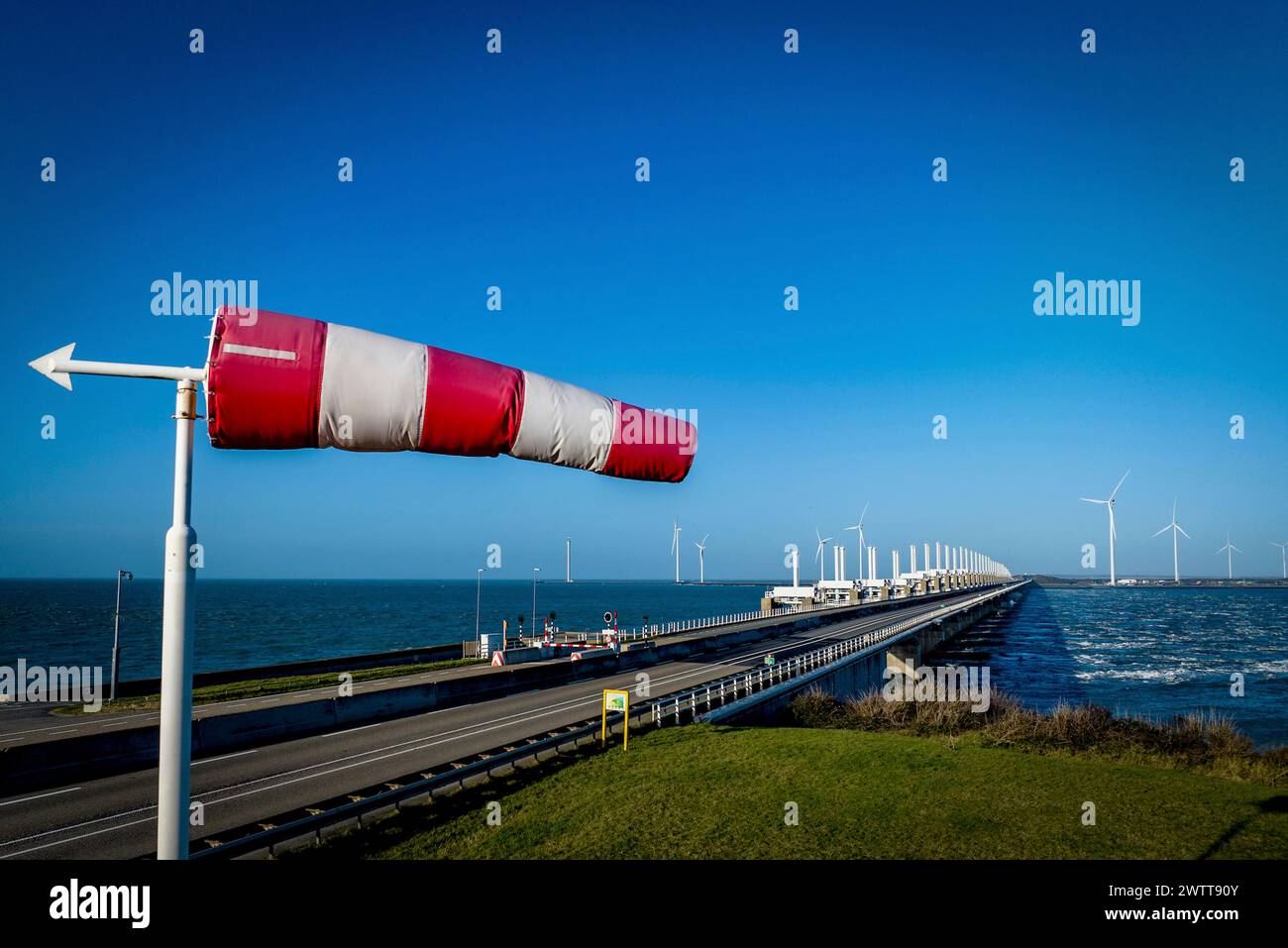 Una calza a vento fluttua vibrante nella brezza accanto a una strada costiera e alle turbine eoliche. Foto Stock