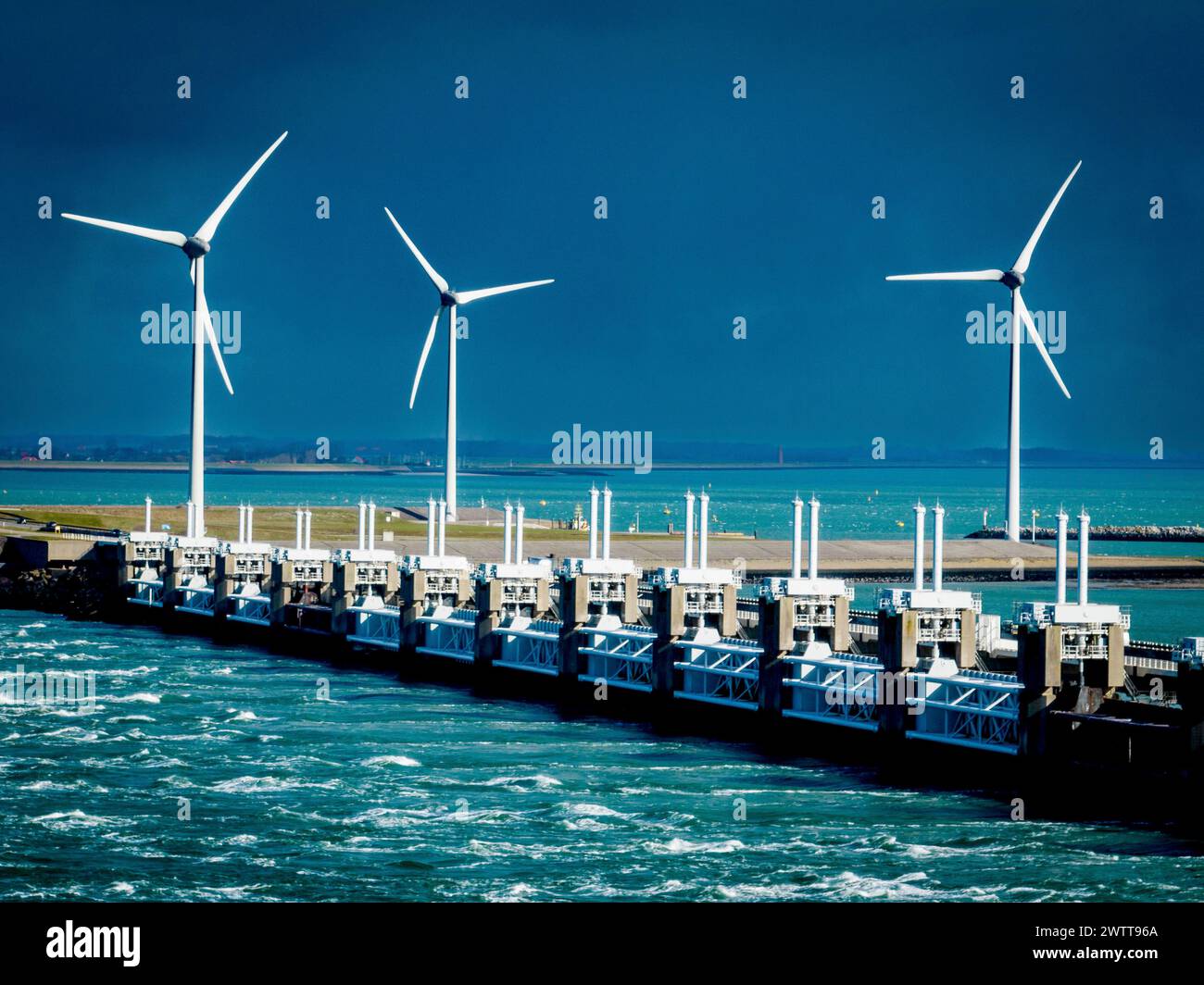 Turbine eoliche che torreggiano su un paesaggio marino tempestoso lungo una barriera costiera. Foto Stock