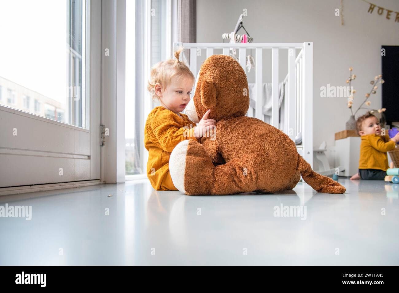 Bambino di colore giallo che abbraccia un grande peluche in una stanza da gioco soleggiata. Foto Stock