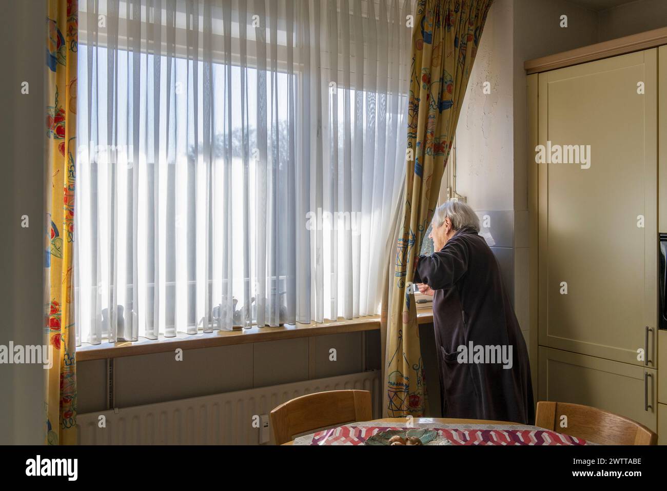 Persona anziana che guarda fuori una finestra soleggiata della cucina in contemplazione. Foto Stock