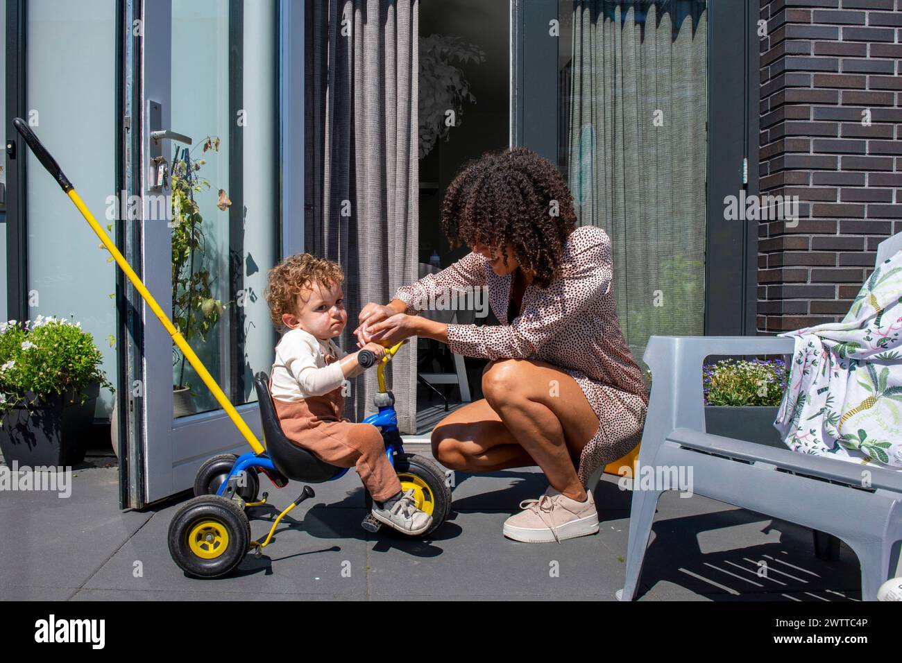 Un bambino che viene amorevolmente assistito da un adulto con la sua bicicletta giocattolo su un patio soleggiato. Foto Stock