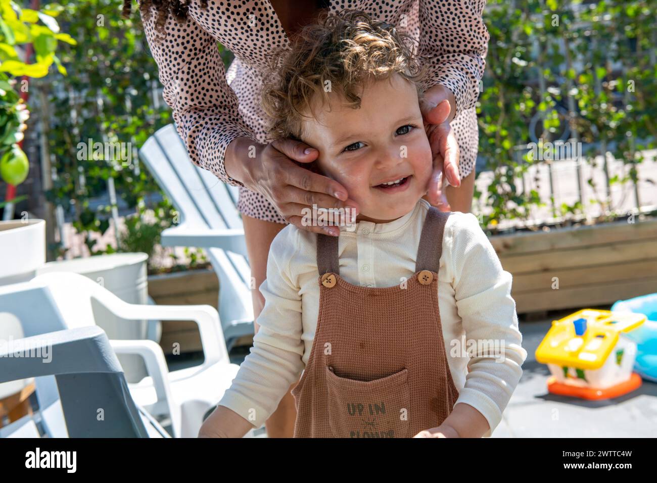 Un bambino allegro si diverte a giocare in un giardino soleggiato con le mani di un adulto che tiene delicatamente il viso del bambino. Foto Stock