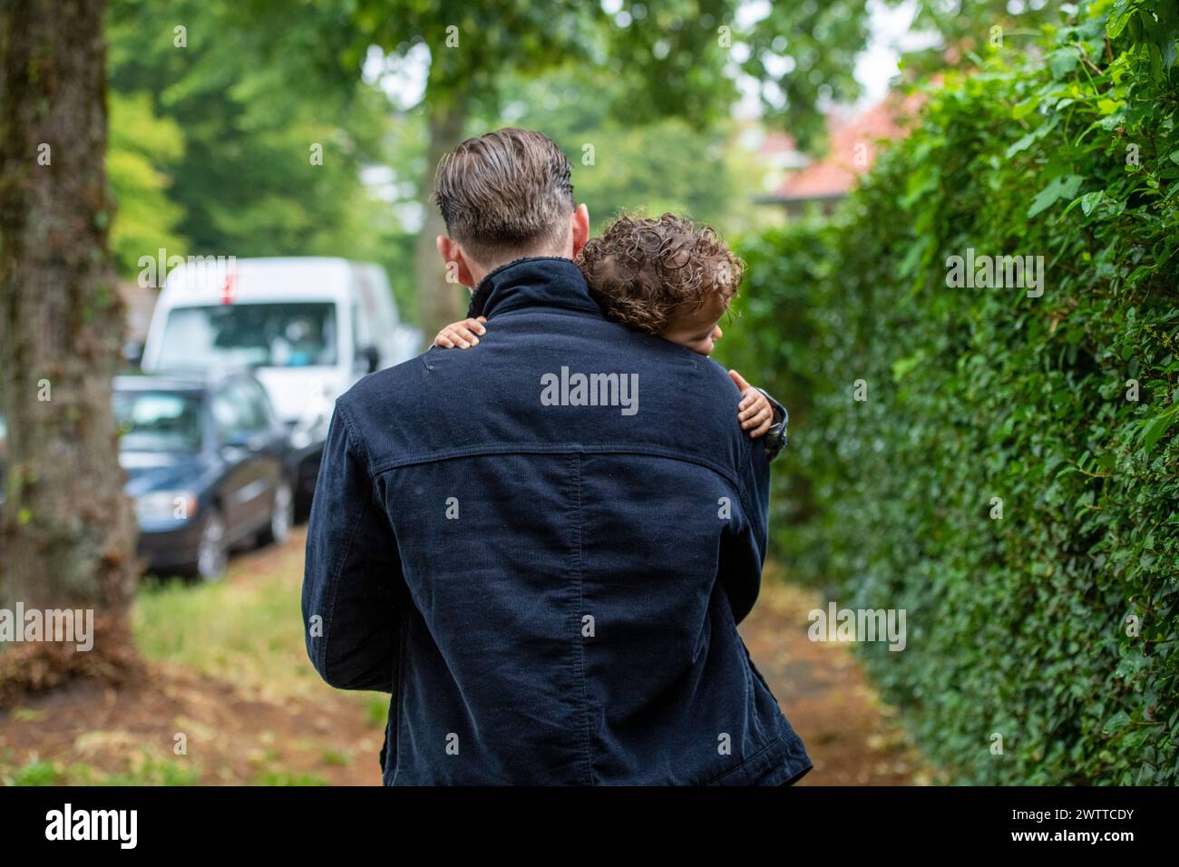 Un momento tenero come padre porta il suo giovane figlio sulle spalle, passeggiando lungo una verdeggiante strada suburbana. Foto Stock