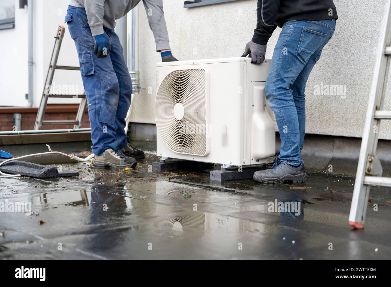 Due tecnici che installano un'unità pompa di calore esterna su una superficie bagnata. Foto Stock