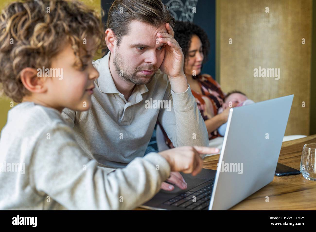 Un bambino fa notare qualcosa su un notebook a un uomo adulto concentrato mentre una donna lavora in background. Foto Stock