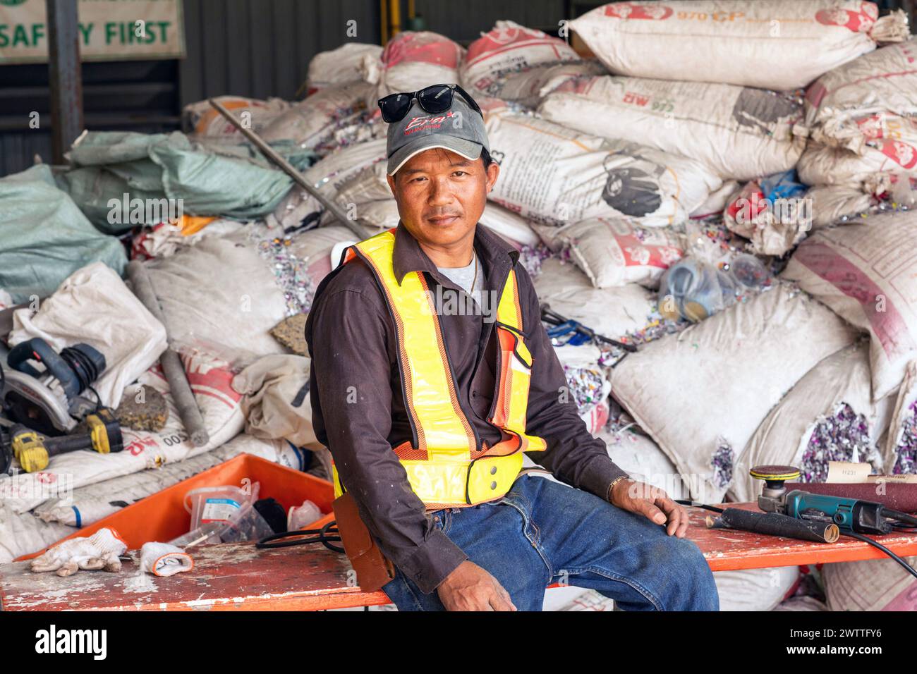 Giovane lavoratore in giubbotto riflettente seduto in mezzo a materiali industriali in una fabbrica di riciclaggio di plastica, in Thailandia Foto Stock