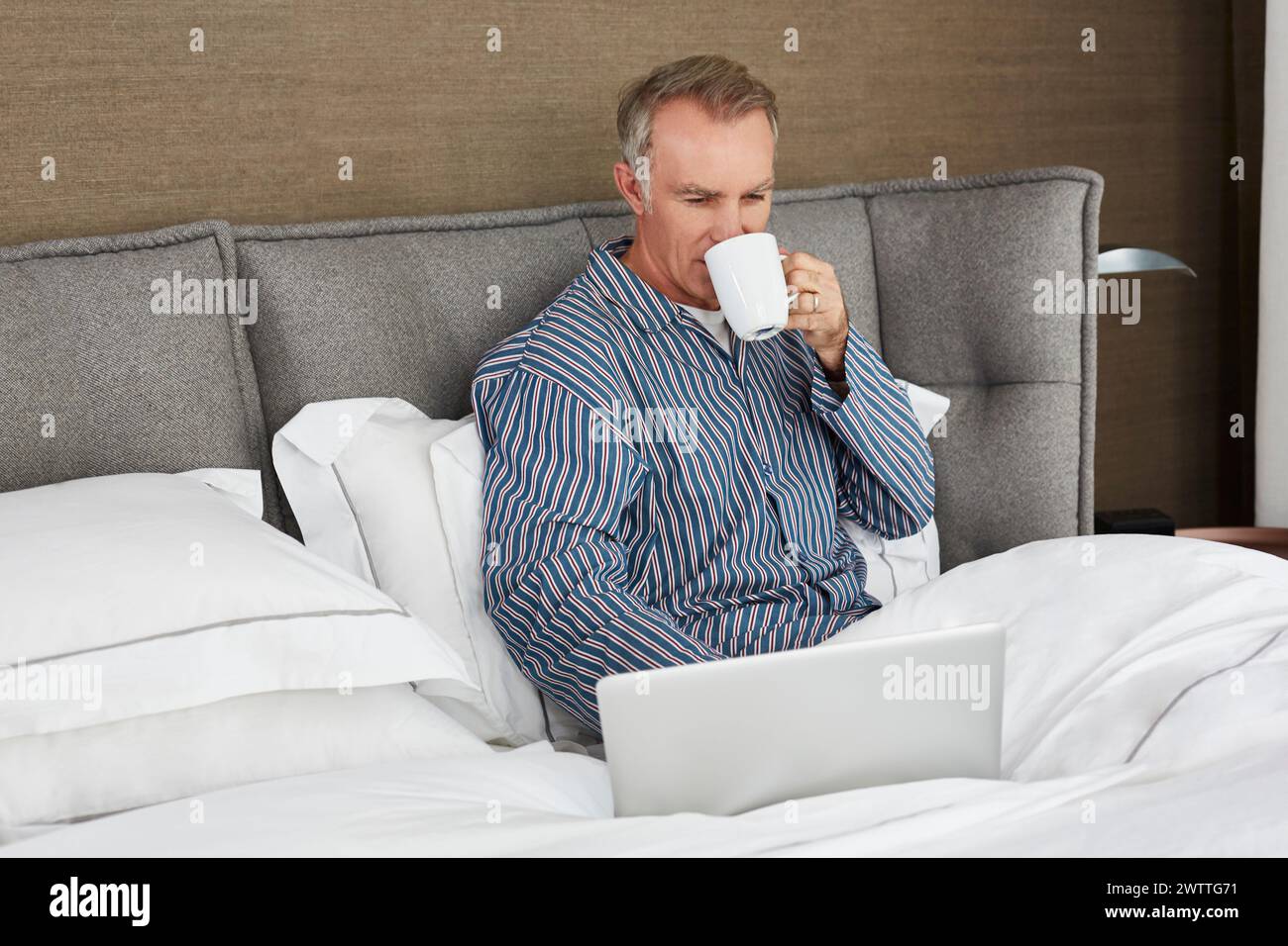 Uomo che lavora al computer portatile a letto mentre beve un caffè Foto Stock