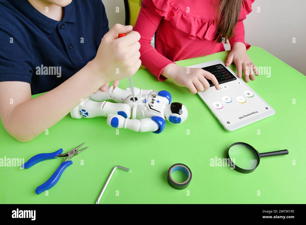 Le mani dei bambini assemblano il robot sul banco di lavoro, avvitando le parti durante la revisione del codice di programmazione sul tablet. Concetto di apprendimento e di formazione pratica Foto Stock