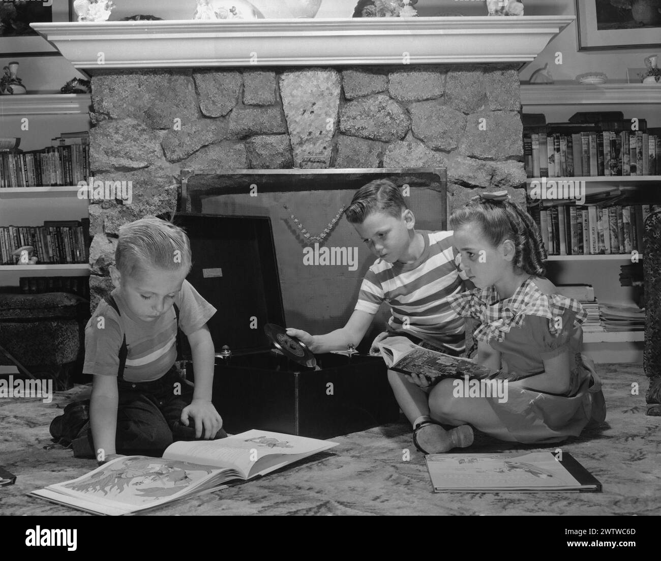 Tre bambini seduti sul piano del soggiorno con libri a piani aperti accanto a un lettore di dischi, che leggono l'audio riprodotto Foto Stock