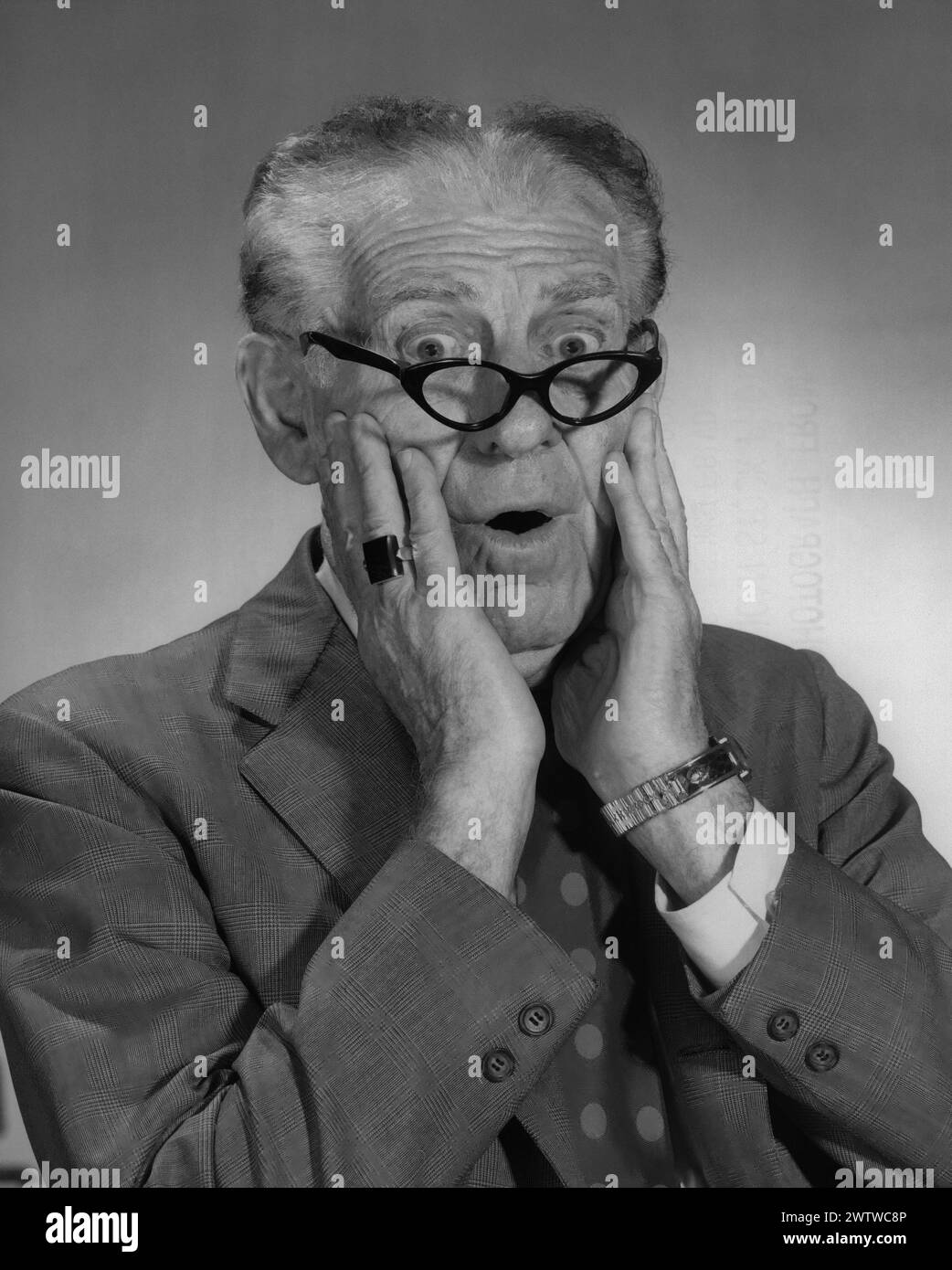 Immagine di un uomo anziano con un abito da lavoro che indossa un orologio, un grande anello e occhiali da lettura che vengono spinti fino al bordo del naso con le mani sul viso, gli occhi spalancati e uno sguardo sorpreso Foto Stock