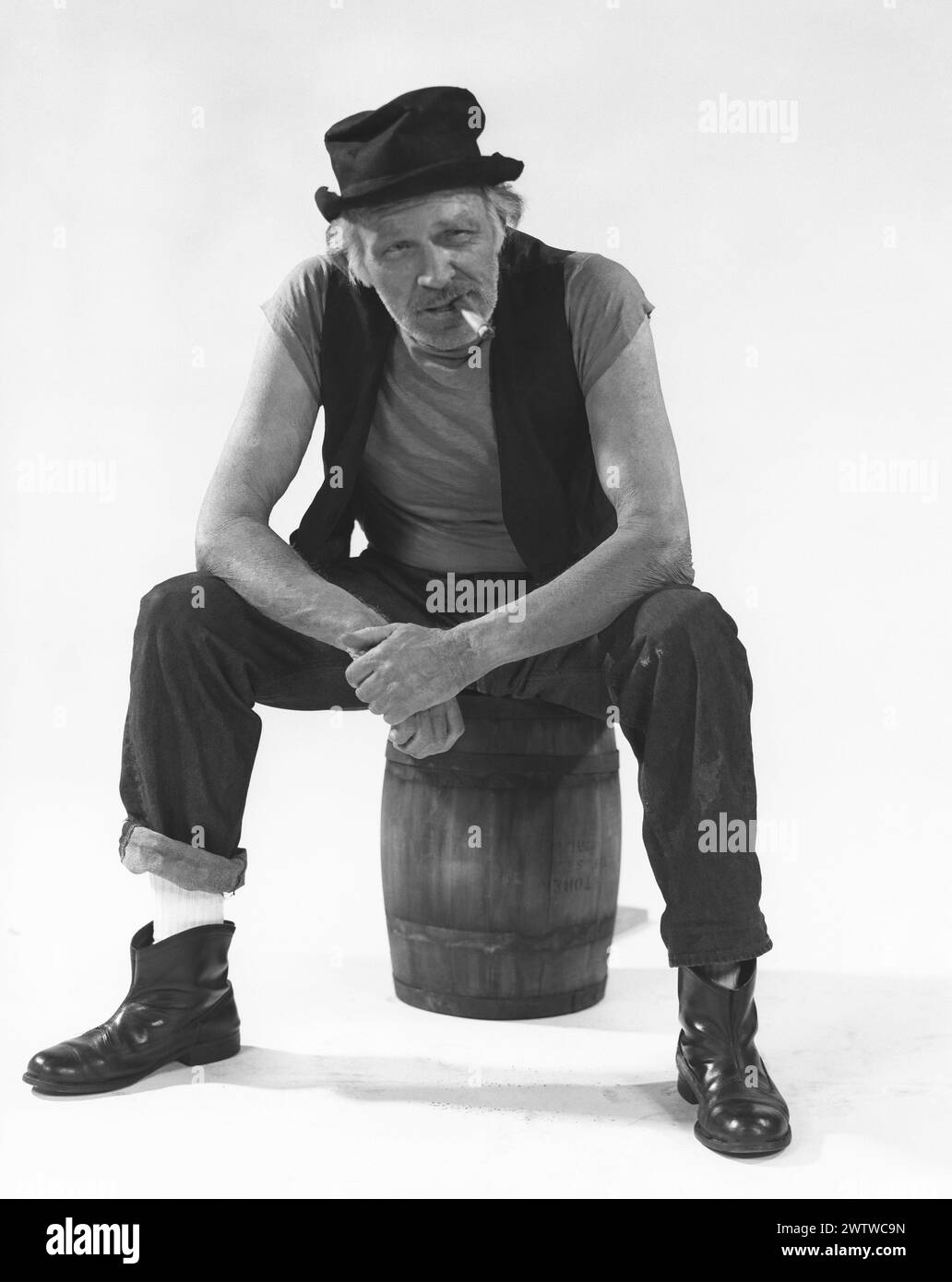 Uomo anziano e dall'aspetto ruvido in un giubbotto e cappello sbriciolato seduto su una botte di legno, le gambe distanti, le mani unite, inclinato in avanti mentre fuma un sigaro Foto Stock