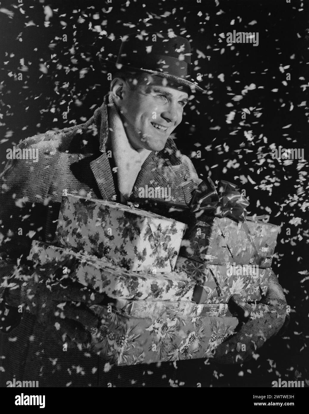 Un uomo di mezza età, raffigurato come una falsa neve che gli gira intorno. Indossa un cappello, un cappotto pesante e guanti e porta con sé una pila di regali di Natale Foto Stock