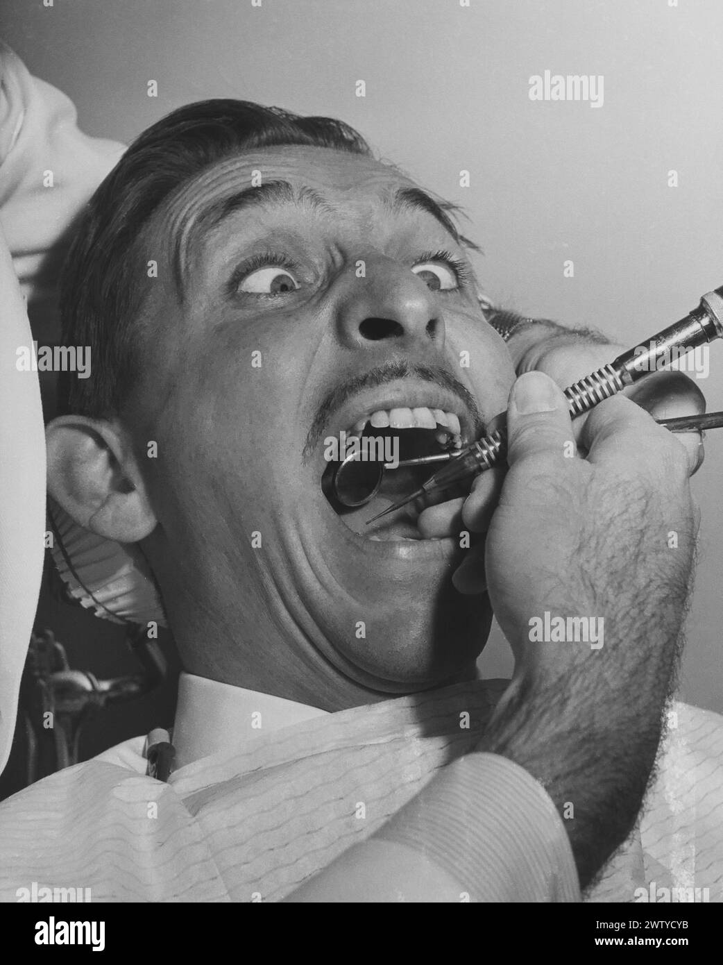 Un uomo di mezza età sembra ansioso mentre siede sulla sedia dentale e sta sottoponendo a una procedura dentale di routine Foto Stock