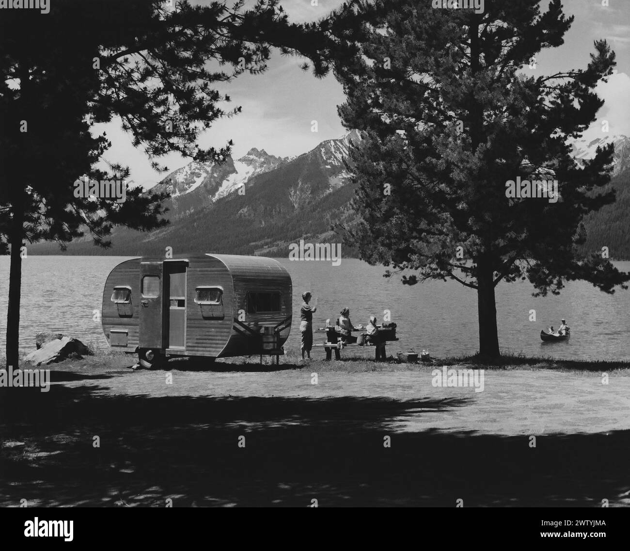 Madre e due bambini seduti a un tavolo da picnic accanto al loro rimorchio che è seduto sulla riva di un lago, una catena montuosa sullo sfondo. Padre e un altro bambino sono in canoa sul lago Foto Stock
