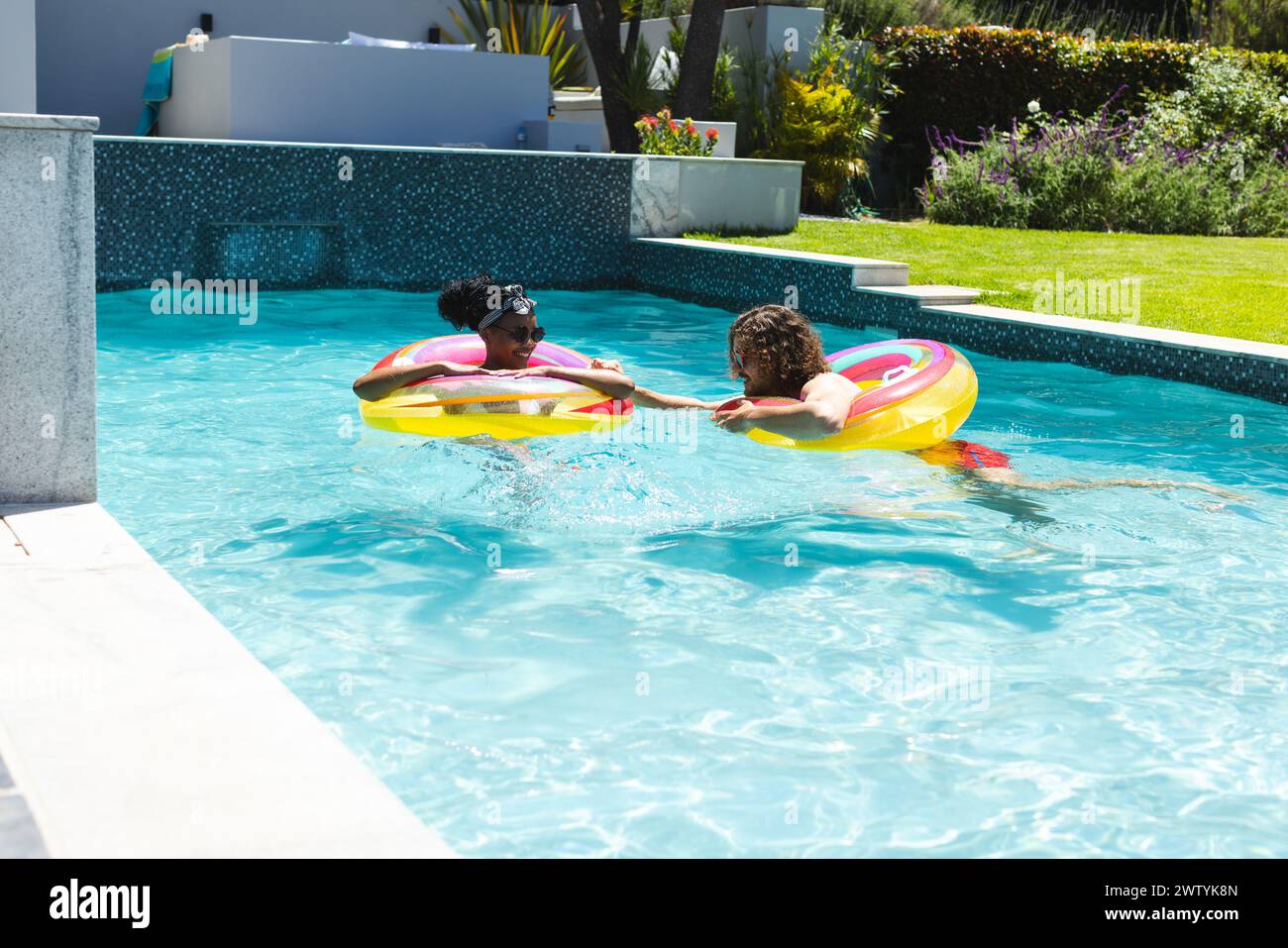 Una coppia eterogenea gode di una giornata di sole in piscina a casa, galleggiando su anelli colorati con spazio fotocopie Foto Stock