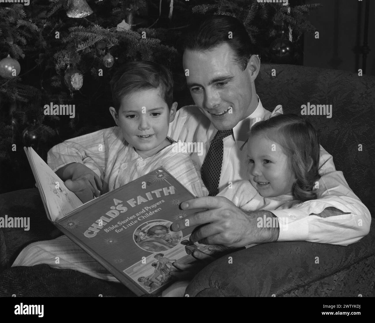 Il giovane papà con una camicia e una cravatta seduto su una poltrona di fronte all'albero di Natale con i suoi due bambini in pigiama seduti in grembo mentre leggono un libro insieme Foto Stock