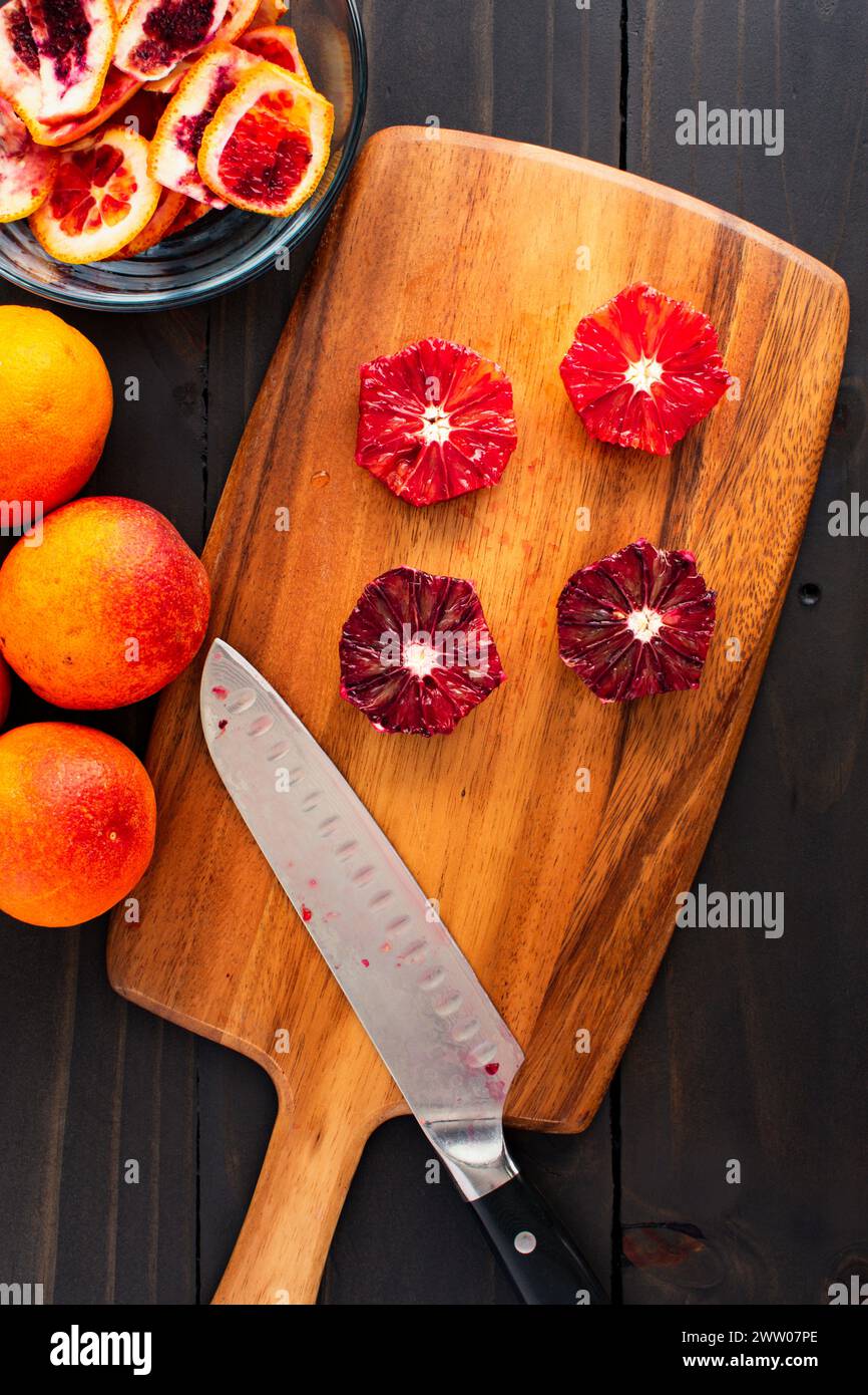 Arance sanguigne sbucciate e deseminate su un tagliere di legno: Arance pelate, tagliate a metà e intere di lamponi con un coltello dello chef e bucce laterali Foto Stock