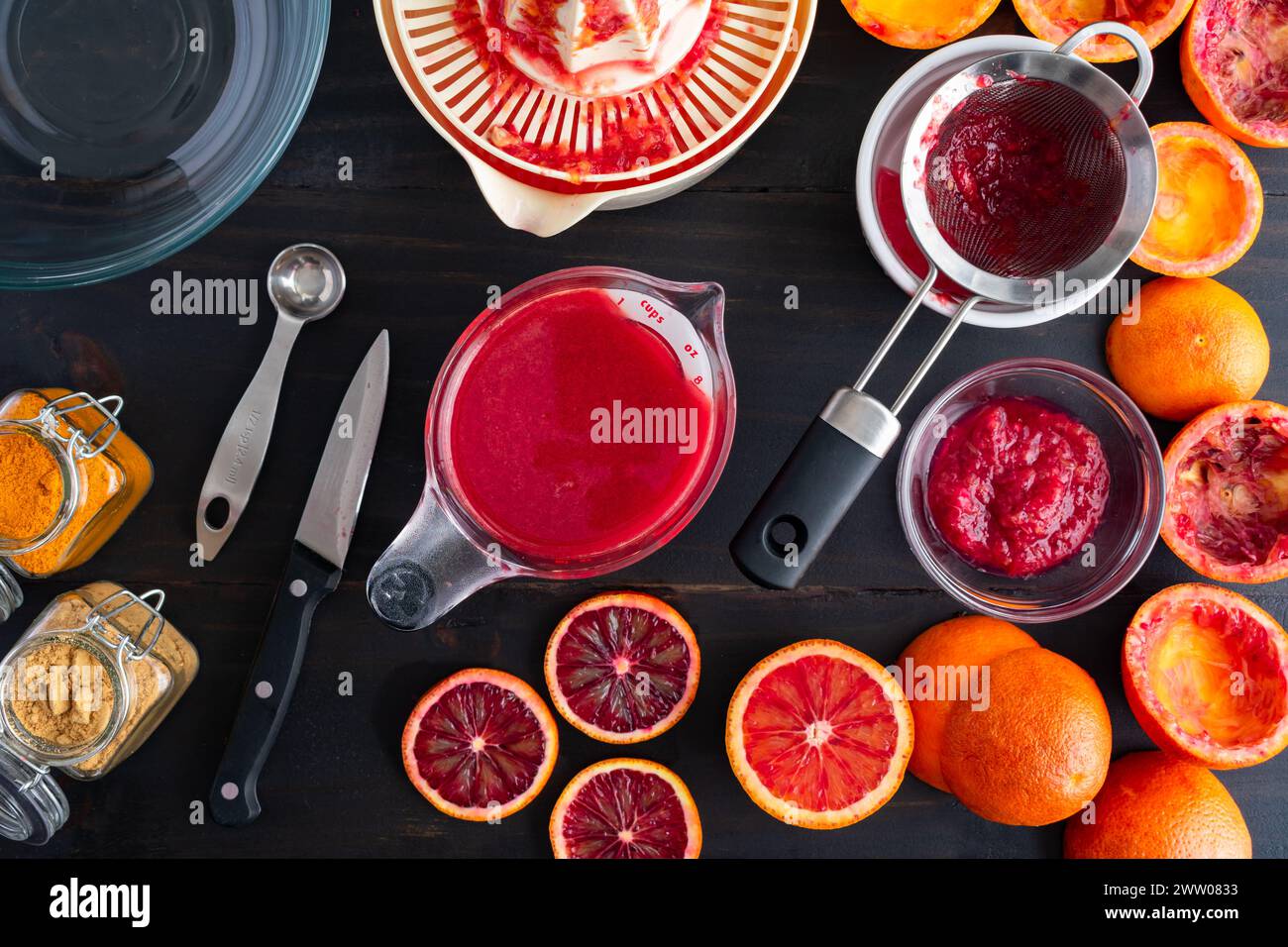 Succo d'arancia ematica in un misurino su un tavolo di legno: Arance ematiche spremute e dimezzate con centrifuga elettrica, succhieruola e altri utensili da cucina Foto Stock