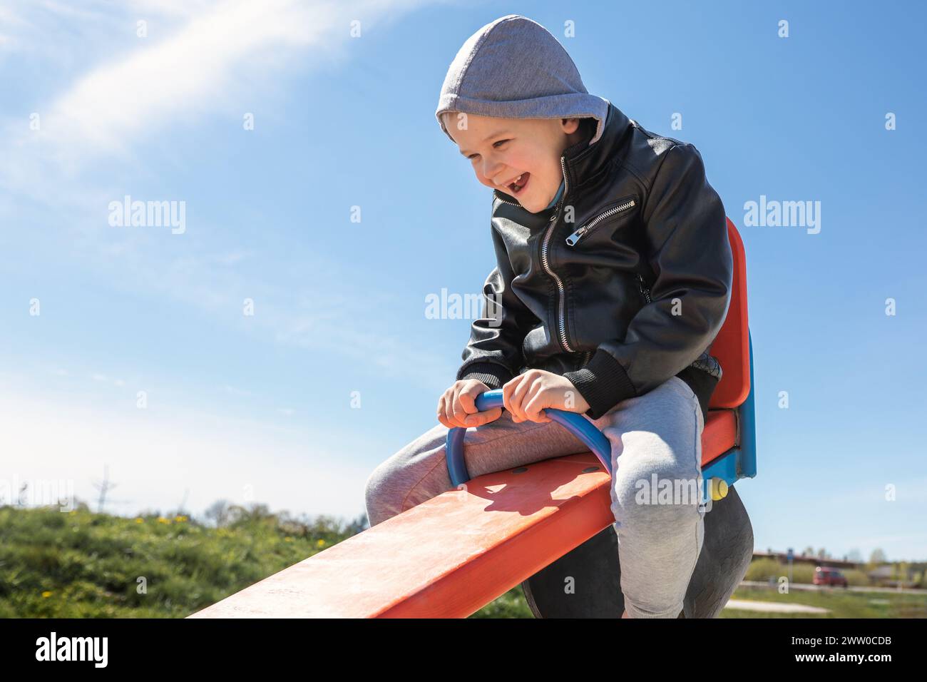 Un bambino si siede e alza su una bilancia rossa all'aperto. Un bambino felice ride, sfondo blu del cielo. Foto Stock