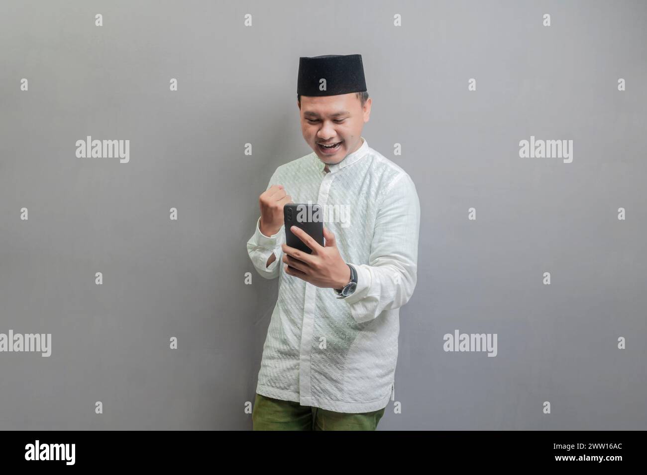 Uomo musulmano asiatico che indossa una camicia koko e un peci con sfumature del mese di digiuno, stringendo il pugno e tenendo in mano il suo smartphone mentre cerca di smartp Foto Stock