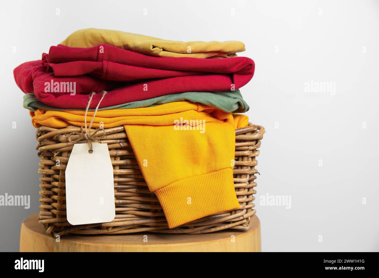 Una pila di capi piegati colorati impilati in un cesto rustico in vimini, completo di un'etichetta vuota pronta per il branding. Foto Stock