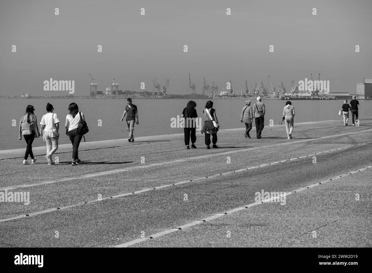 Molte persone con abbigliamento informale, camminano sul molo di cemento vicino al mare Egeo. Bianco e nero, colori. Monocromatico. 10. 14. 2023 Salonicco Foto Stock