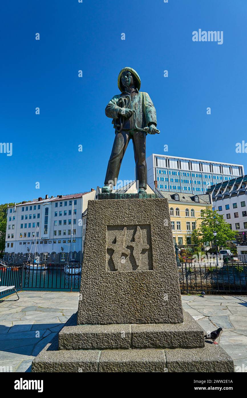 Alesund, Norvegia - 06 07 2022: Statua in bronzo di Skarungen fisher boy nel centro della città di Alesund lungo il porto con case colorate sullo sfondo, Foto Stock