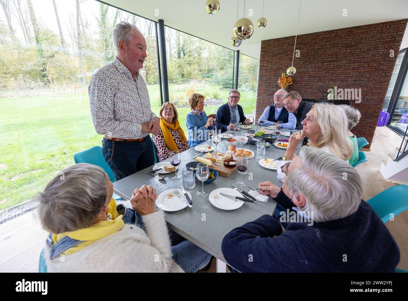 In una sala piena di luce con ampie vedute del giardino, un uomo si erge per rivolgersi agli ospiti seduti per un brunch informale. L'ambiente moderno e arioso e' completato da una parete d'accento in mattoni rustici, che evoca una miscela di raffinatezza e calore. Uomo in piedi e rivolgendosi alla famiglia in un casual Home Brunch. Foto di alta qualità Foto Stock