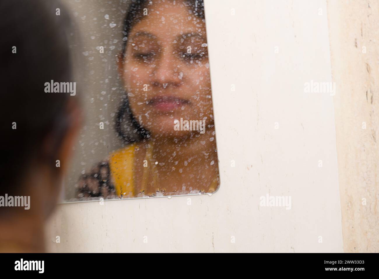 Una giovane donna pensiva sta in un bagno a vapore, guardando il suo riflesso nello specchio Foto Stock