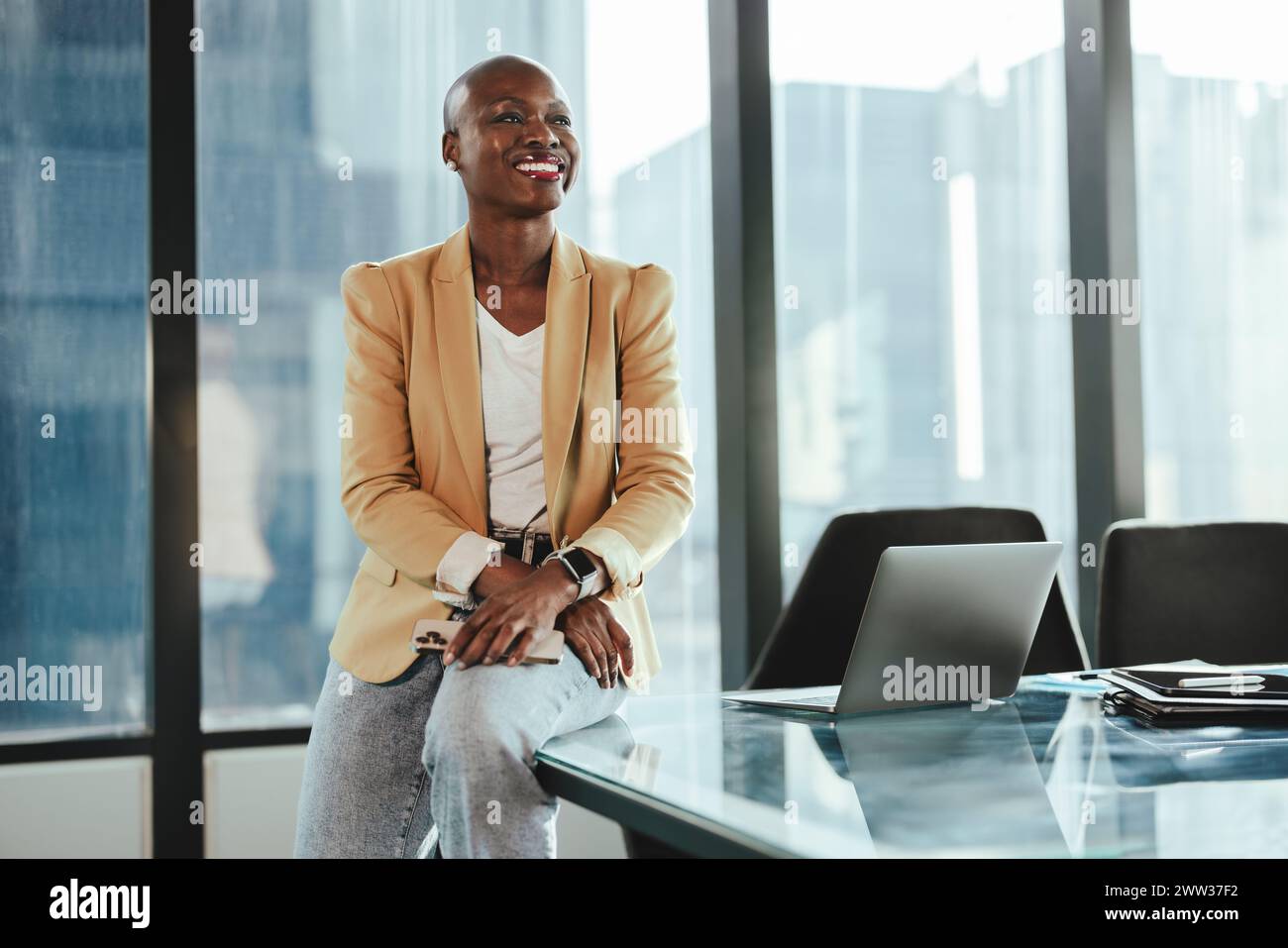 Donna d'affari africana sicura di sé in un ufficio, seduta a un tavolo. Lei è felice e di successo, sorride mentre guarda lontano. La sua professionalità e la sua ent Foto Stock