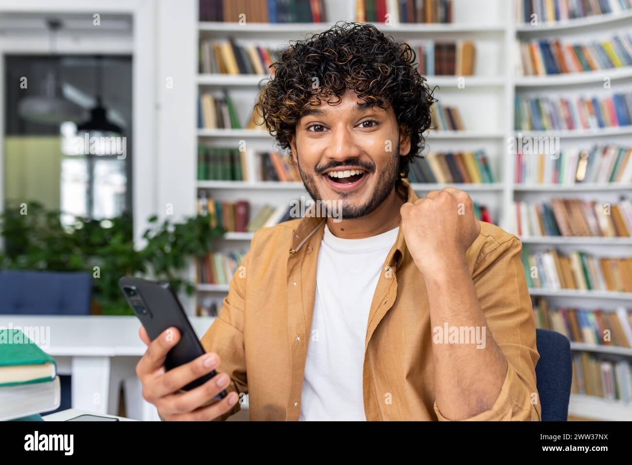Gioioso giovane adulto in una camicia casual che celebra il successo mentre tiene in mano un telefono cellulare, con sfondo da libreria. Foto Stock