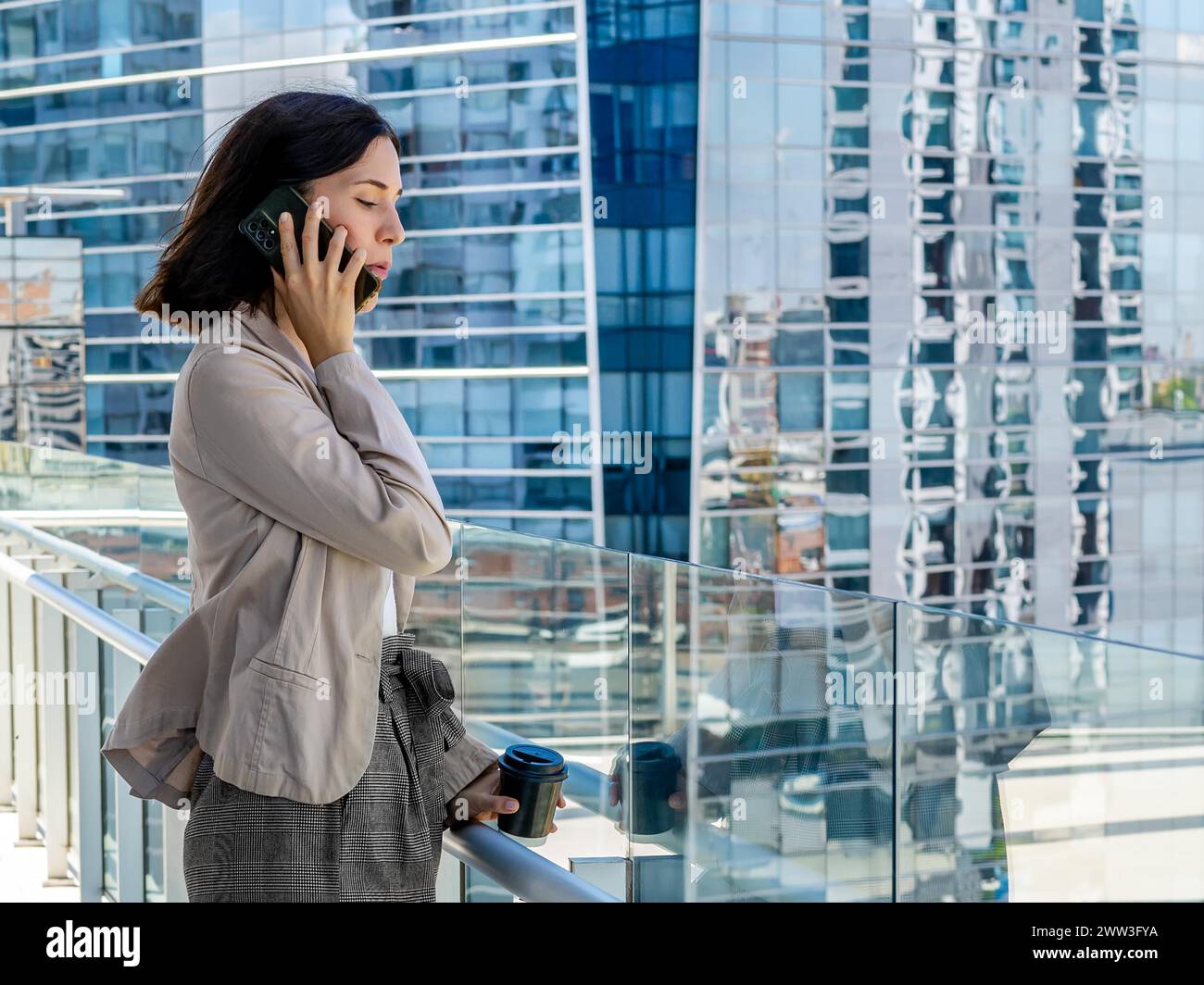 Una donna è in piedi su un balcone, tenendo una tazza di caffè di carta e parlando al cellulare durante la pausa di ufficio Foto Stock