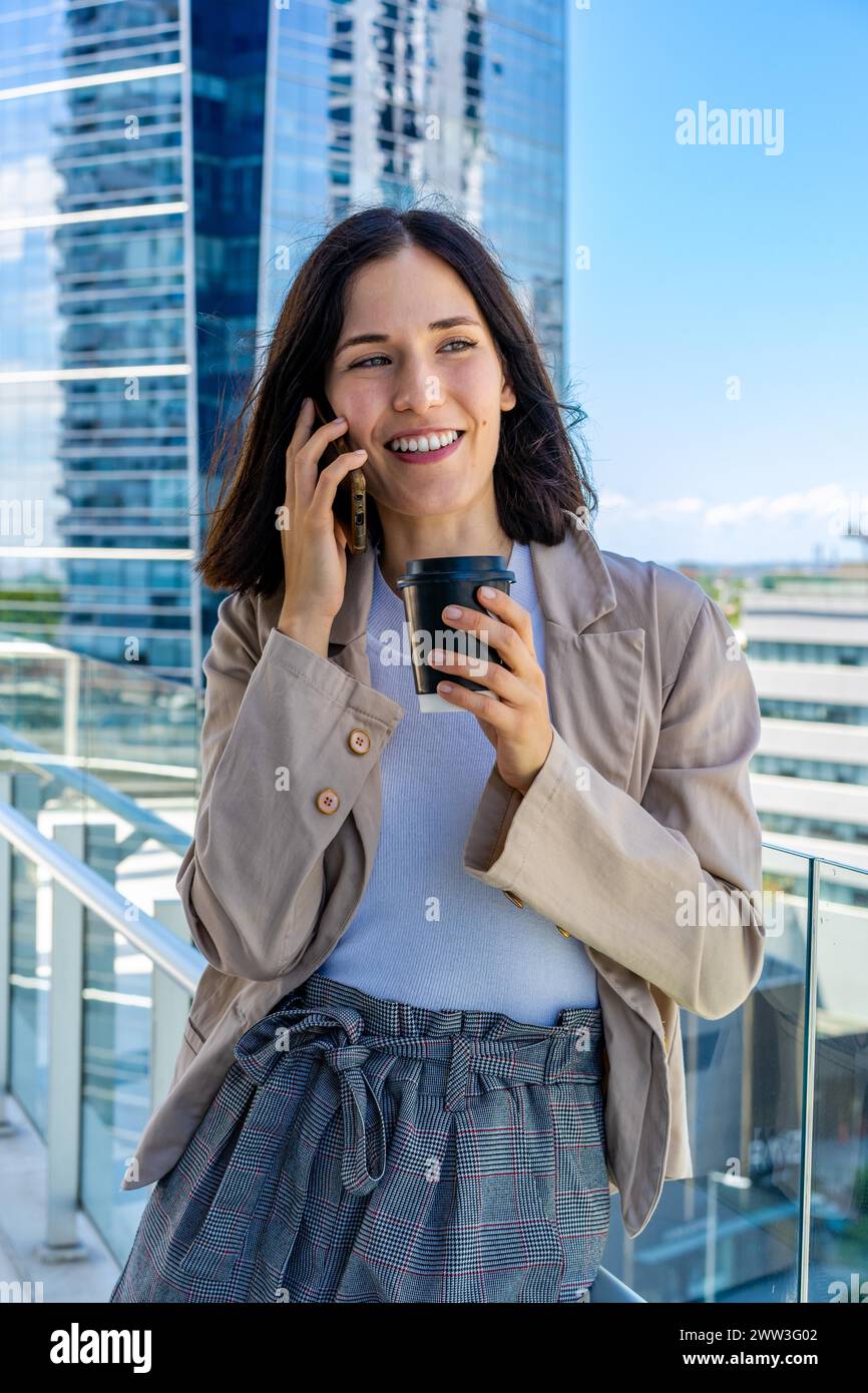 Una donna sta in piedi su un balcone e sta parlando al cellulare durante la pausa del suo ufficio. Ha una tazza di caffè di carta in mano Foto Stock