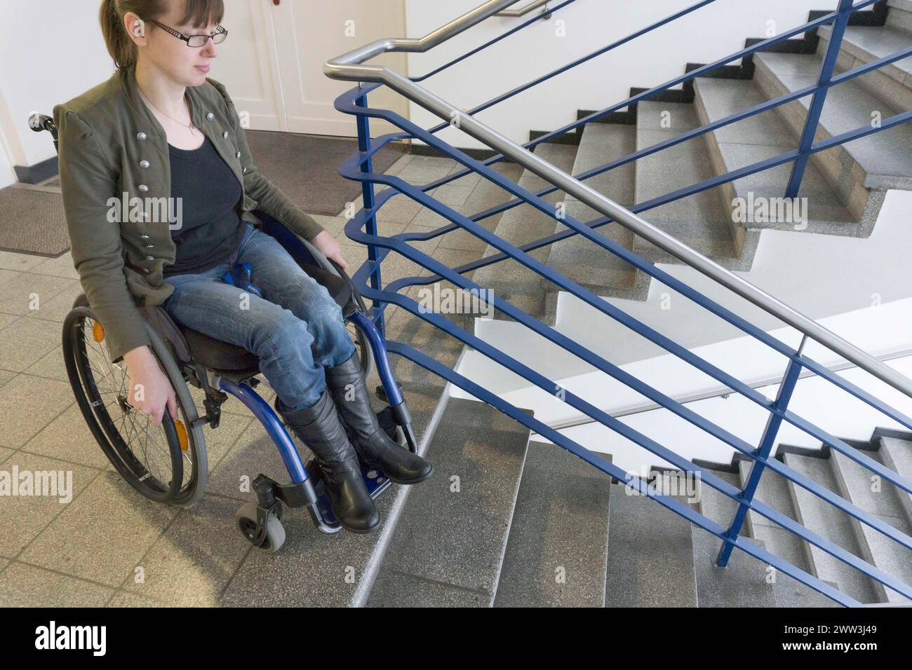 Una giovane donna in sedia a rotelle sta con la sua sedia a rotelle in una scala di un edificio in cerca di accesso senza barriere, Berlino, 18/01/2017 Foto Stock