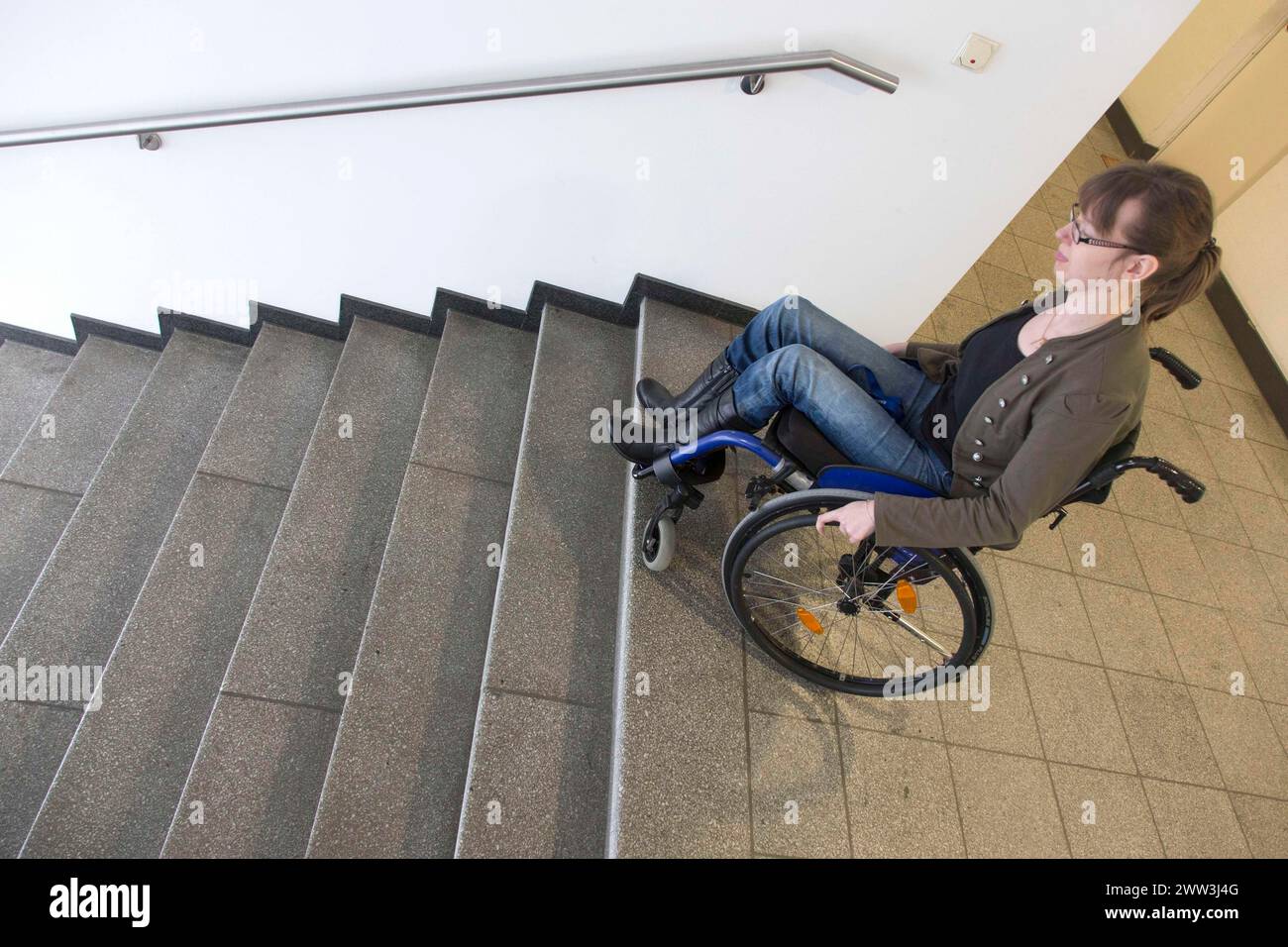 Una giovane donna in sedia a rotelle sta con la sua sedia a rotelle in una scala di un edificio in cerca di accesso senza barriere, Berlino, 18/01/2017 Foto Stock