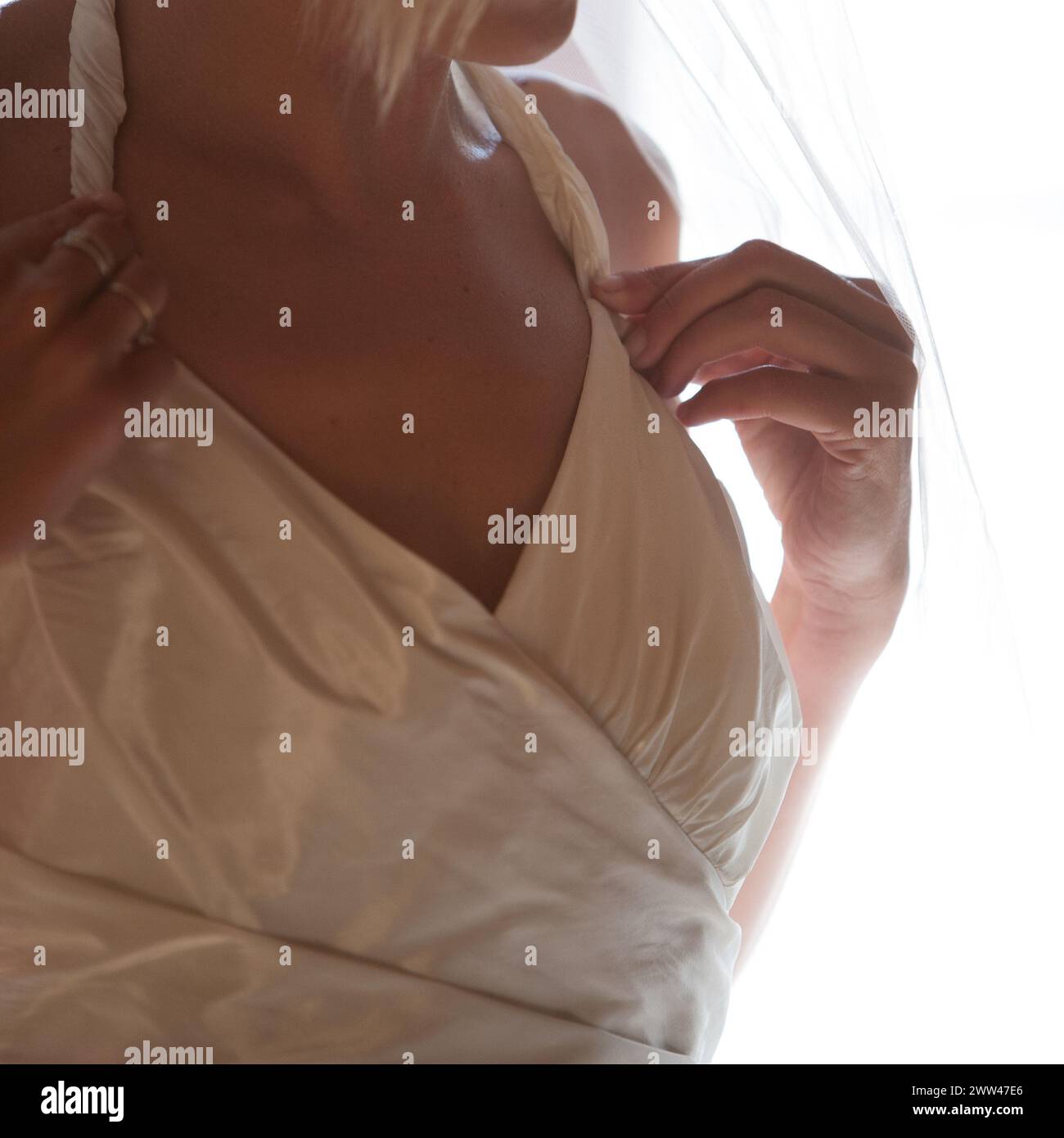 Questa fotografia intima si concentra sulla silhouette di una sposa, catturando la grazia e il fermento mentre regola il suo abito. La morbida retroilluminazione crea un delicato gioco di luci e ombre, mettendo in risalto i dettagli raffinati del suo abbigliamento da sposa. Eleganza nuziale: Uno sguardo di grazia nell'abbigliamento da sposa. Foto di alta qualità Foto Stock