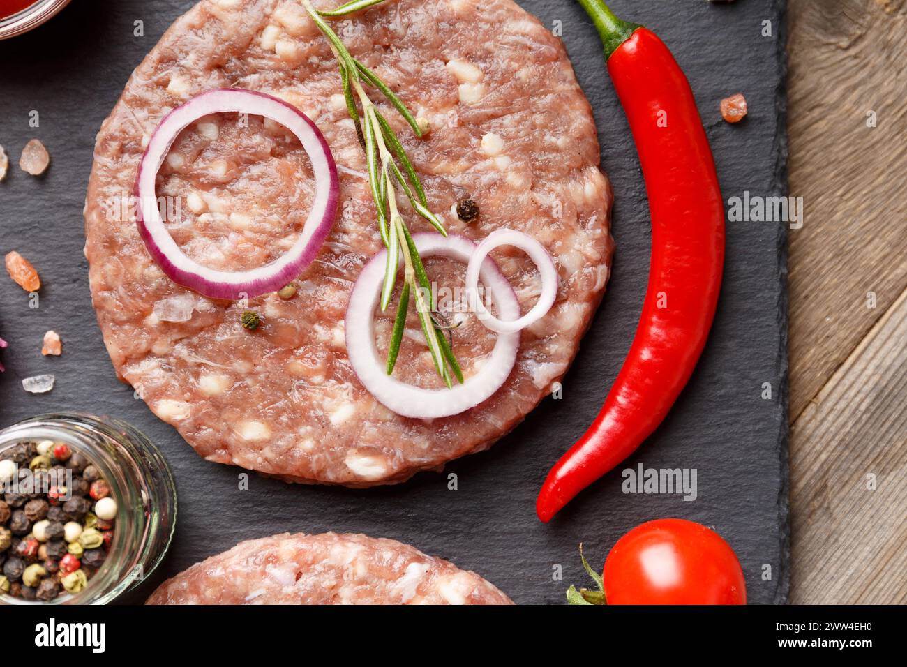 Hamburger crudo di manzo macinato, rosmarino, verdure e spezie su un piatto di pietra nera su uno sfondo di legno, vista dall'alto, primo piano. Foto Stock