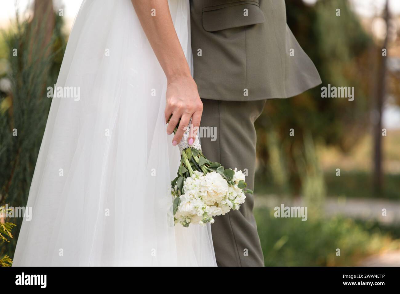 Un paio di sposi novelli nel parco, lo sposo abbraccia la sposa, la sposa regge un bouquet di nozze bianco tra le mani, primo piano Foto Stock