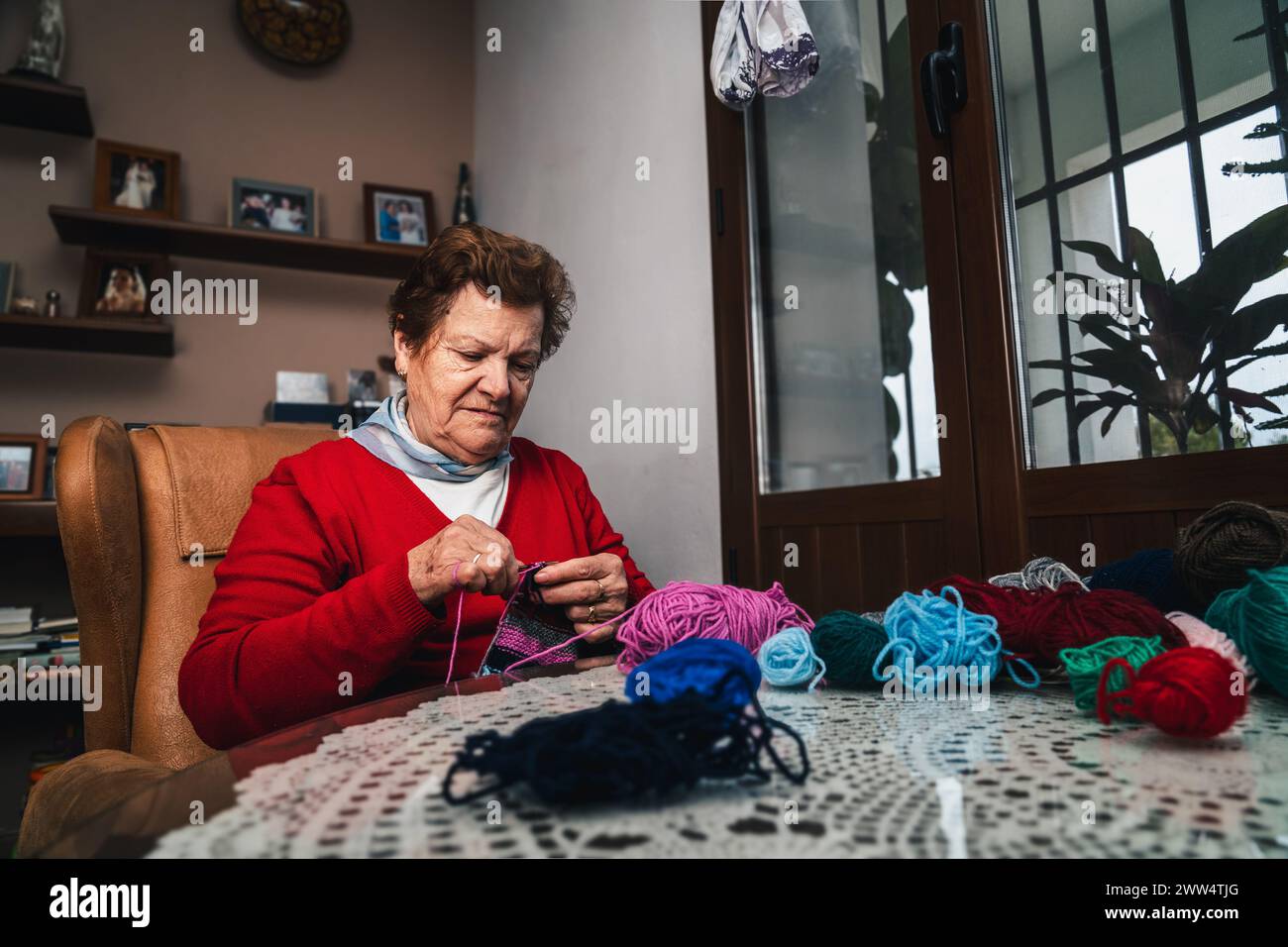 donna anziana che cucisce a mano con aghi e lana, lavora a maglia un capo di abbigliamento di lana nel soggiorno di casa seduto sul divano Foto Stock