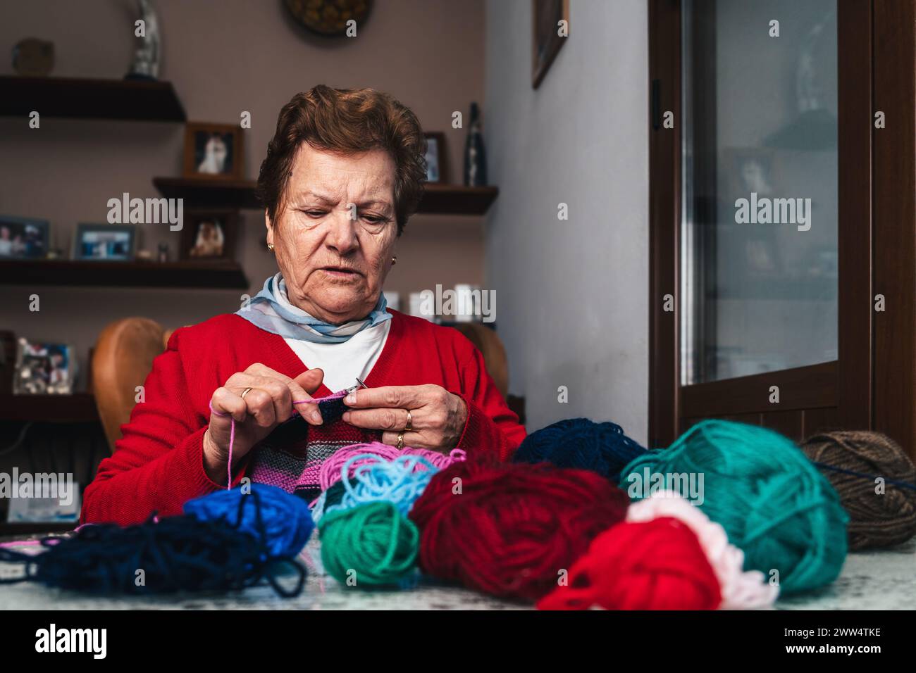 ritratto di una donna anziana carina e felice che cucisce un pezzo di indumento di lana con aghi e diverse palle di lana colorata Foto Stock