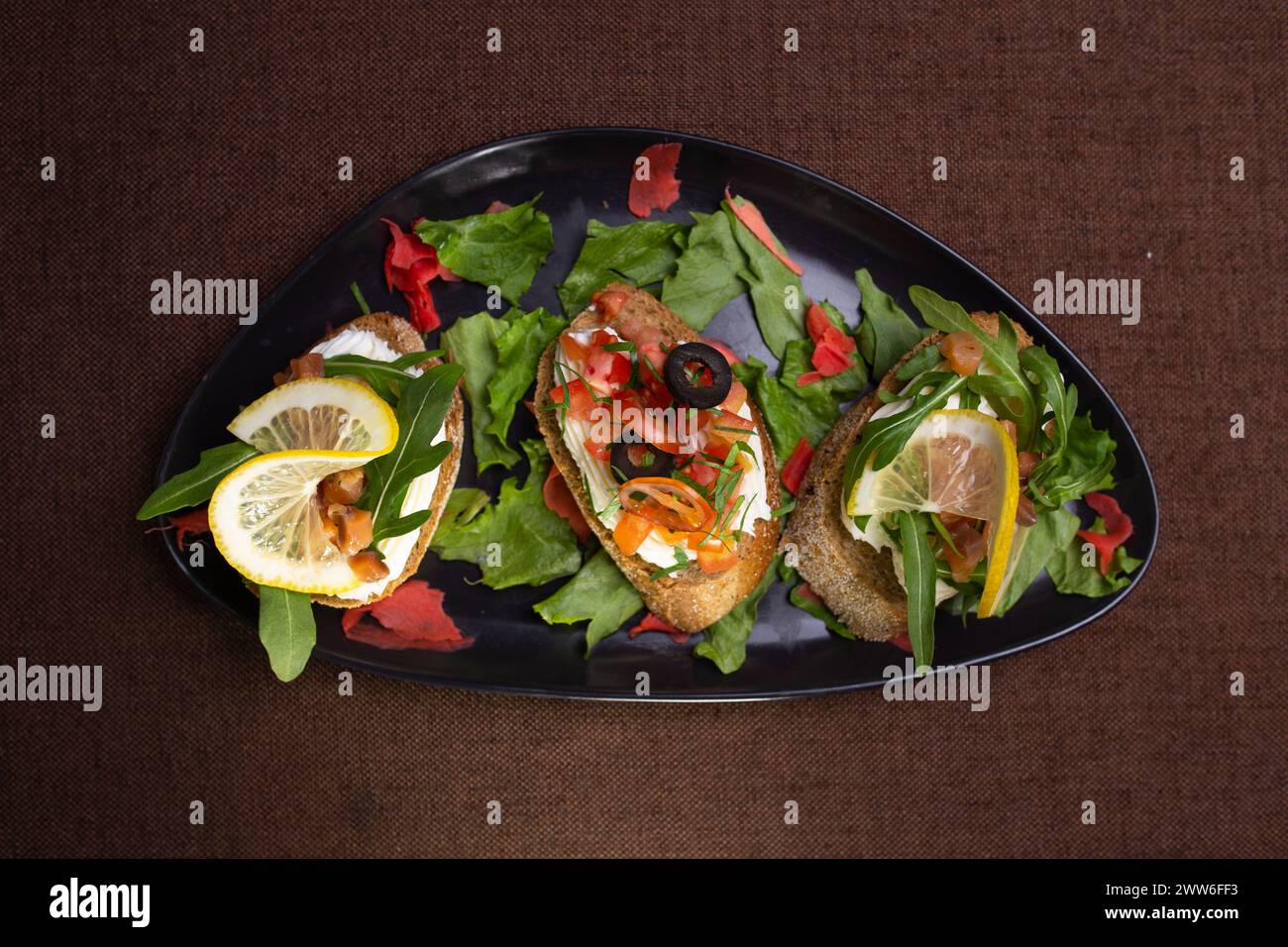 Una varietà di bruschette con formaggio spalmabile, pomodori, olive, limone su un piatto nero, con lattuga e contorno di zenzero sottaceto rosso Foto Stock