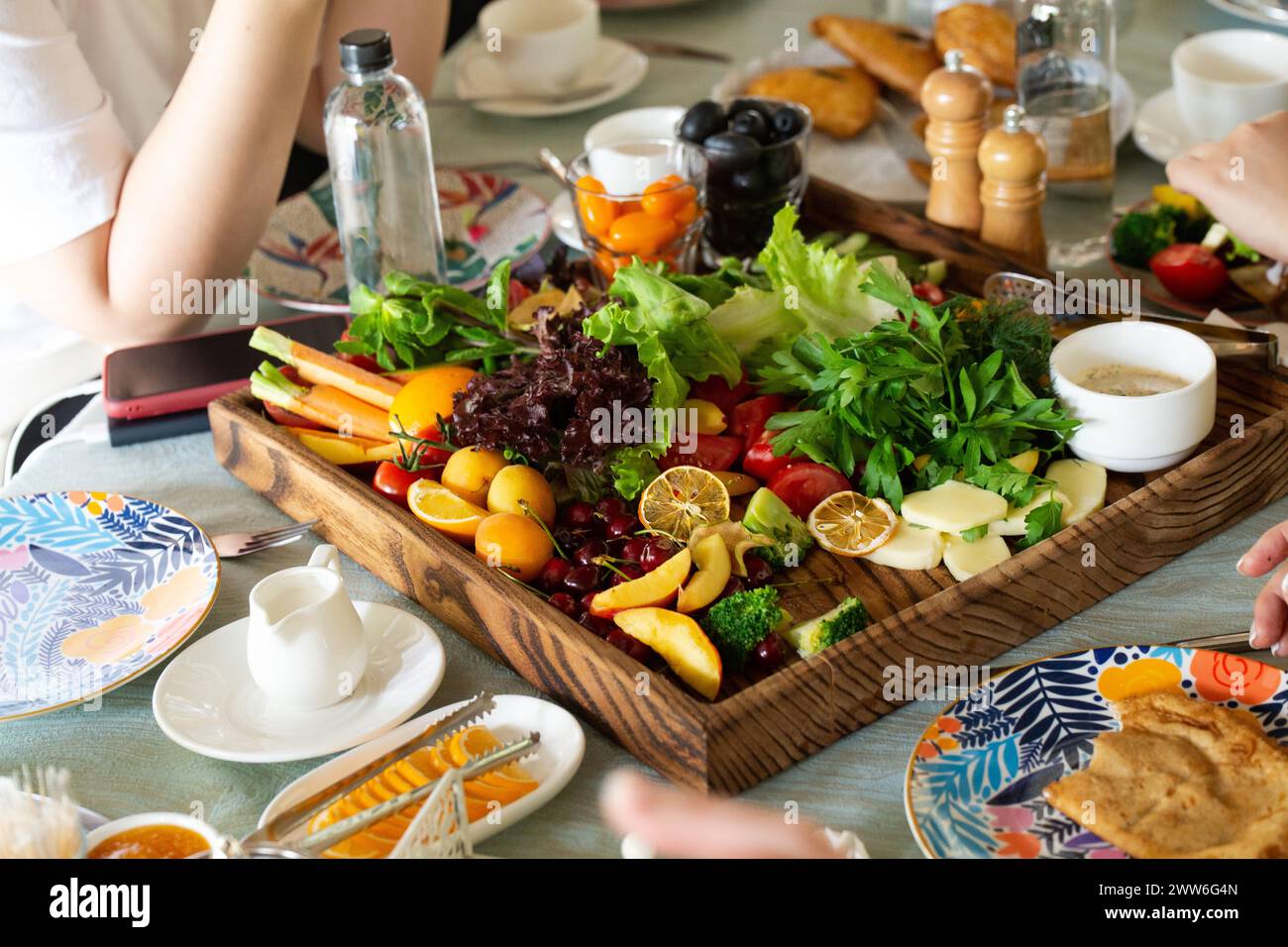 Verdura fresca, frutta, pane su un tavolo di legno con piatti colorati, bicchieri e caraffa d'acqua. Sfondo sfocato della sala da pranzo. Foto Stock