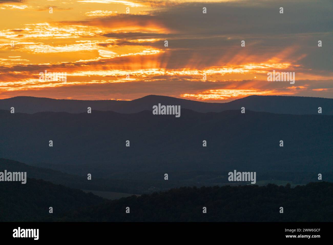 Raggi di luce arancione si sparpagliano sotto nuvole stratificate sopra una fascia di montagne blu e un primo piano boscoso scuro nello Shenandoah National Park, Virginia. Foto Stock