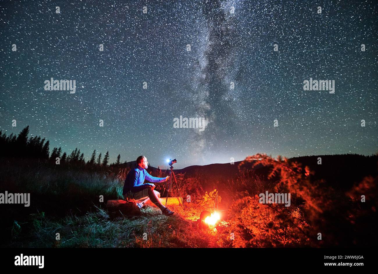 Giovane uomo che scatta foto di cielo stellato in montagna. Fotografo maschio seduto su una macchina fotografica di legno e messa a fuoco. Uomo con macchina fotografica seduta accanto al fuoco del campo. Foto Stock