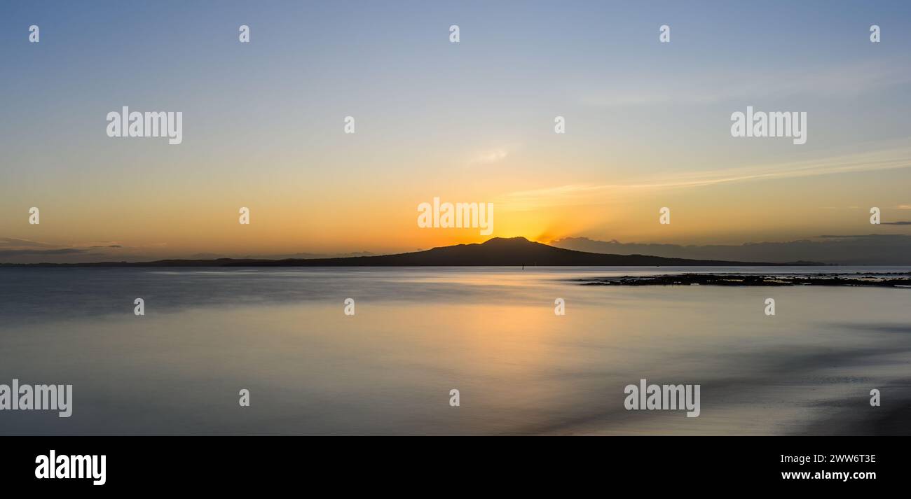 Bagliore dorato in cima all'isola di Rangitoto mentre il sole sorge sopra l'orizzonte. Milford Beach. Auckland. Foto Stock