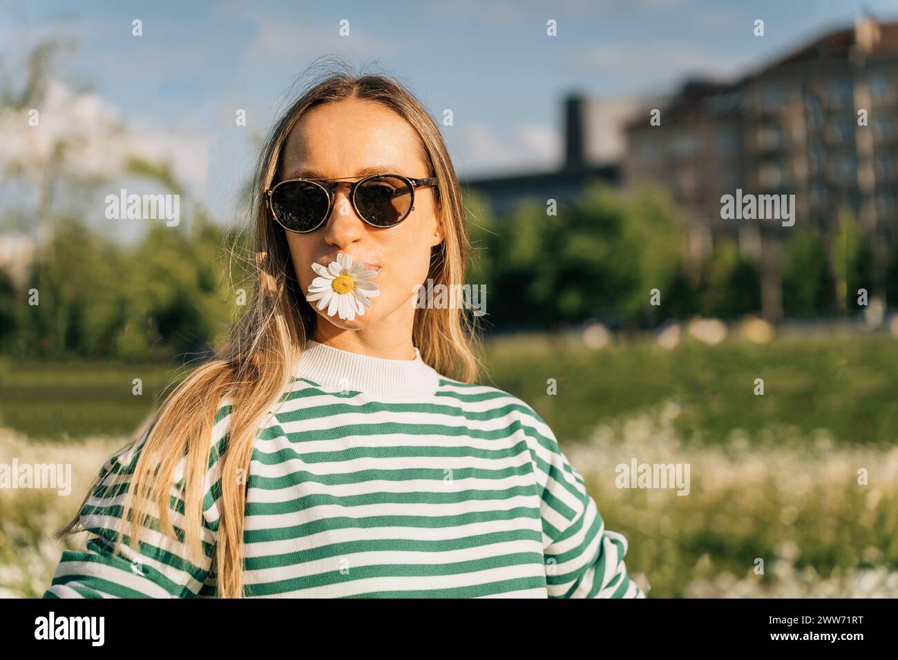 Ritratto di una donna dai capelli lunghi in occhiali da sole con un fiore di camomilla in bocca Foto Stock