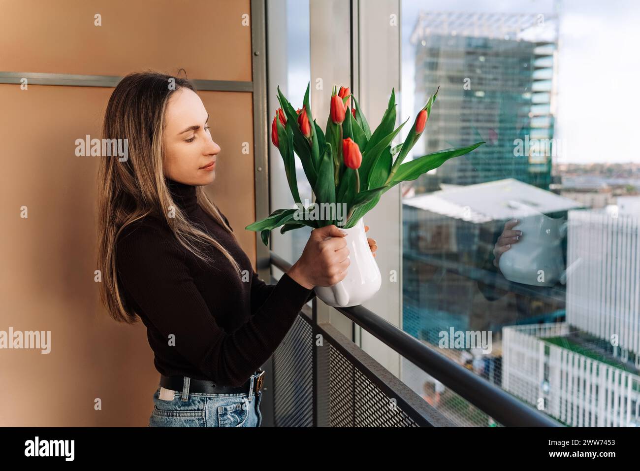 Donna dai capelli lunghi che guarda il bouquet di tulipani nel vaso in piedi vicino alla finestra Foto Stock