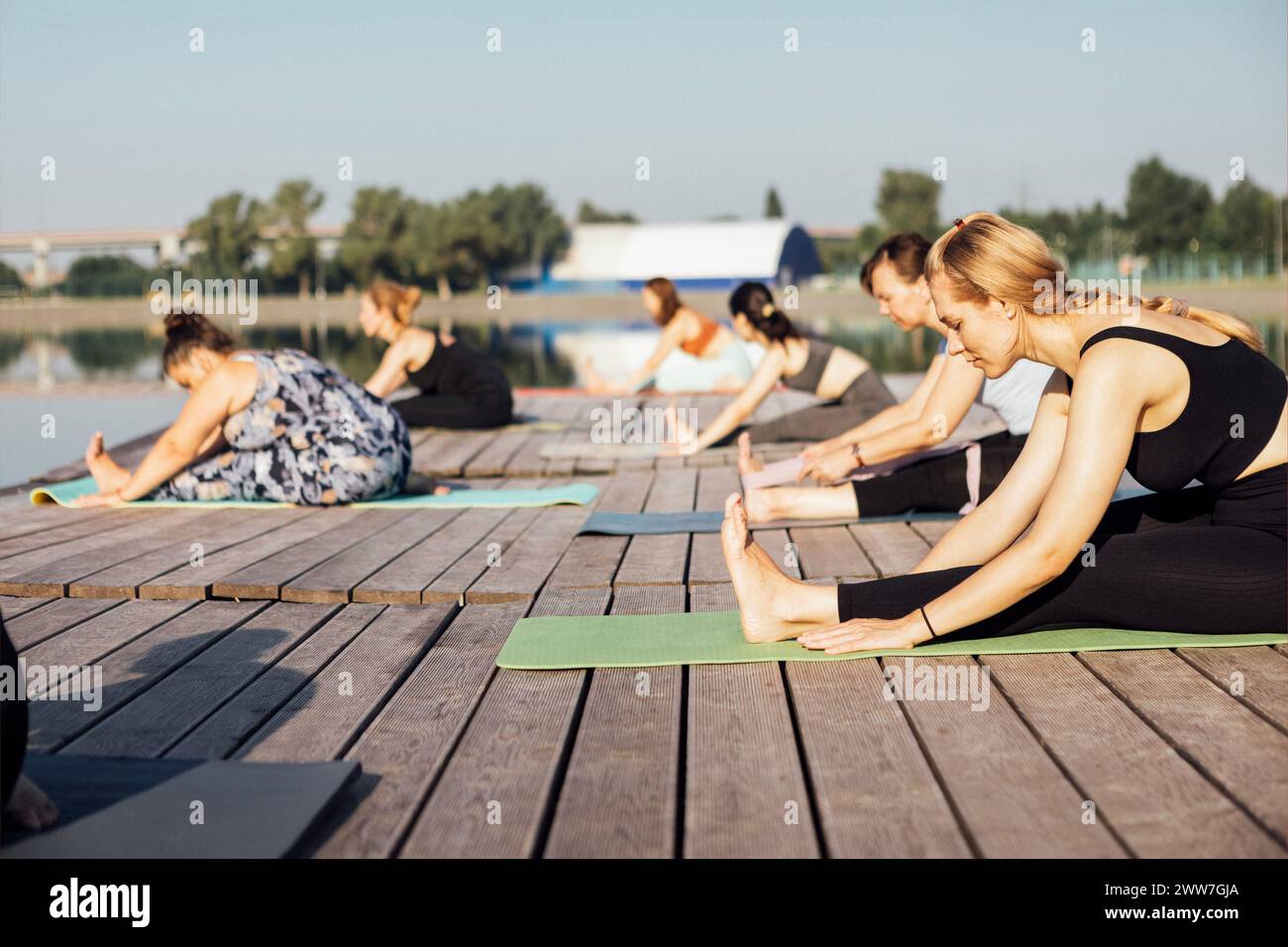 Lezione di yoga mattutina con istruttore vicino al fiume in città. Le giovani donne si piegano mentre si siedono sui tappetini in un molo di legno. Un gruppo di ragazze multietniche sono in agguato Foto Stock
